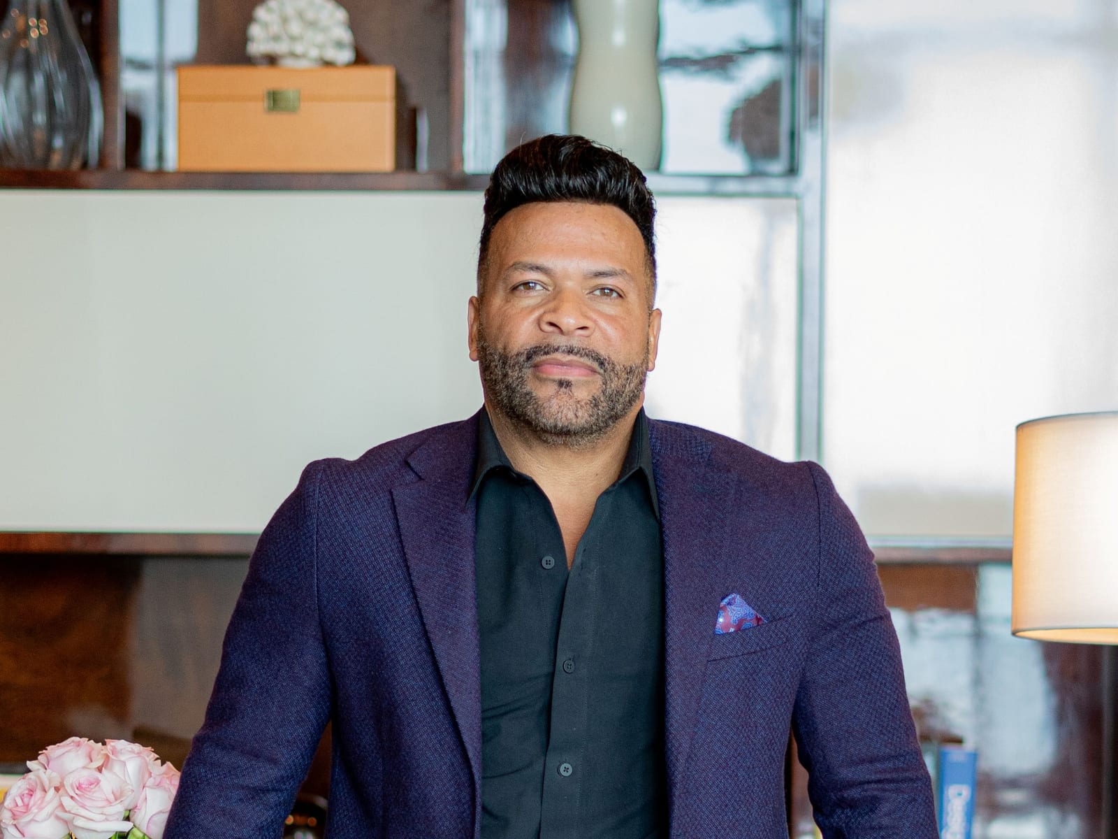 Chris Cole in a dark shirt and patterned navy blazer, leaning against a wooden table in an office with shelving and a vase of pale roses behind him.